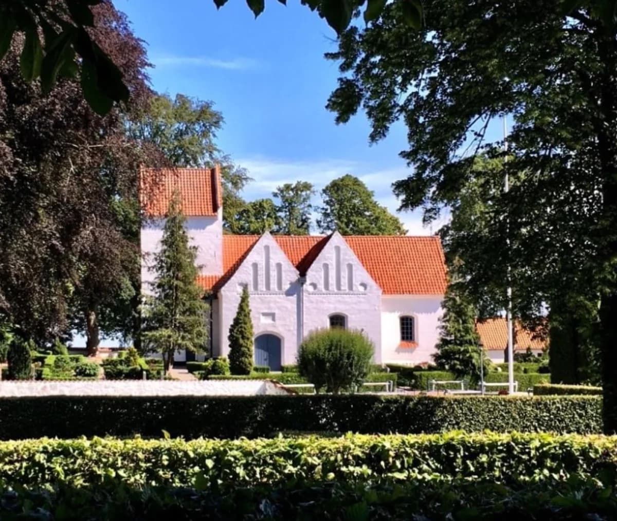 A white church with a red roof surrounded by trees and hedges.