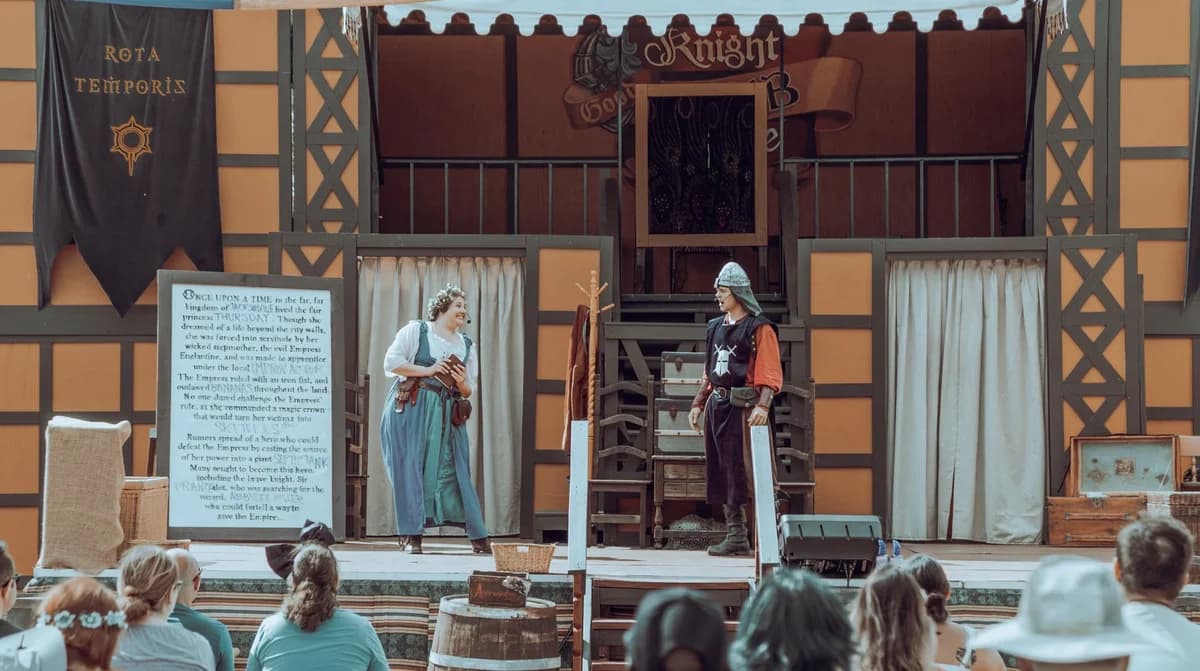 Two costumed actors perform on an outdoor medieval-style stage while spectators watch.