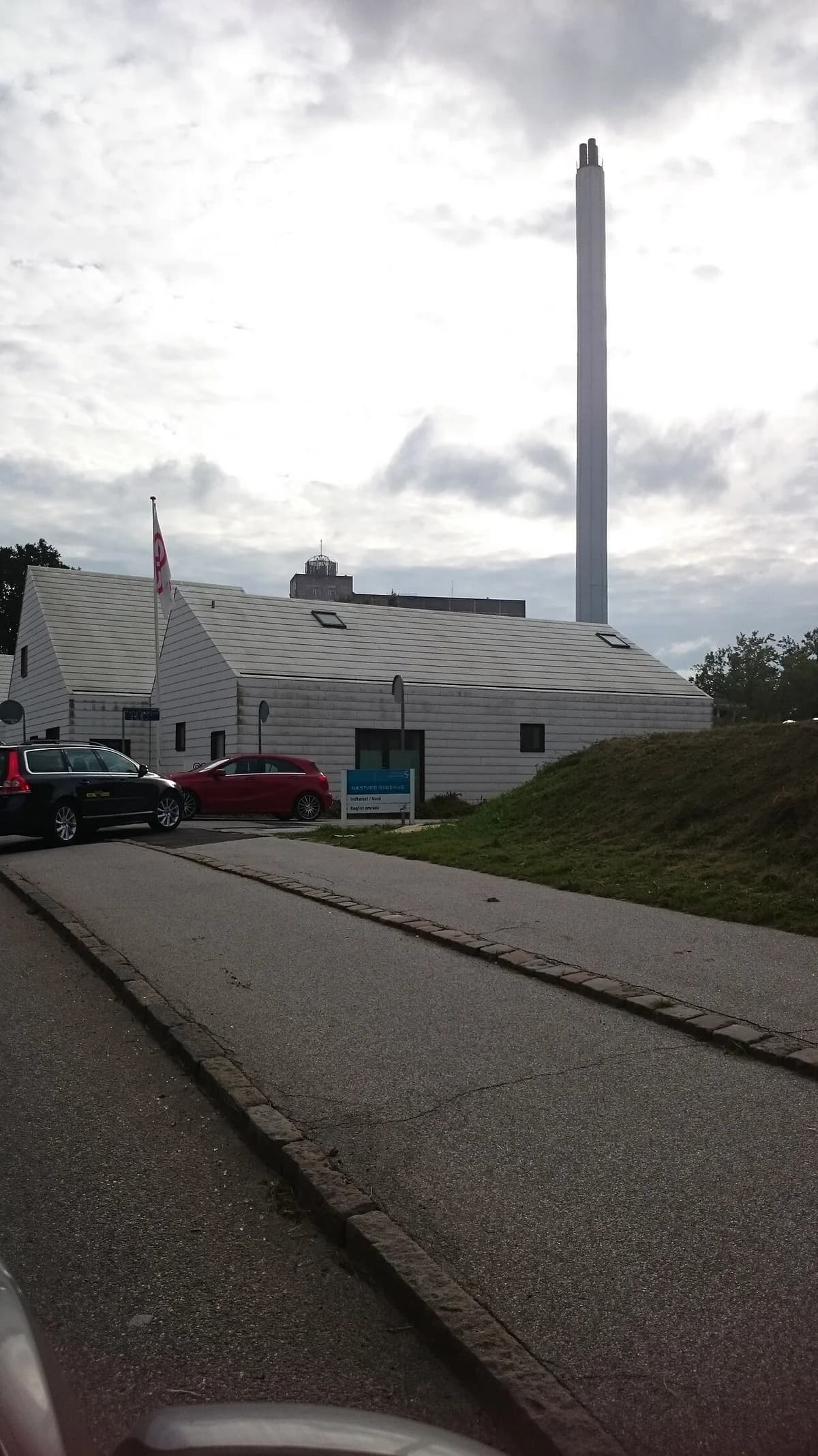 Exterior of the Cancer House at Næstved Hospital with parked cars and a tall chimney.