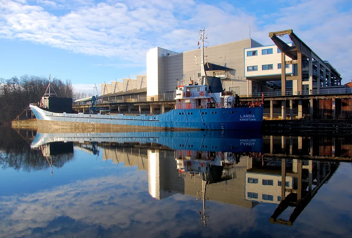 Blue cargo ship Landia docked by industrial waterfront with reflection
